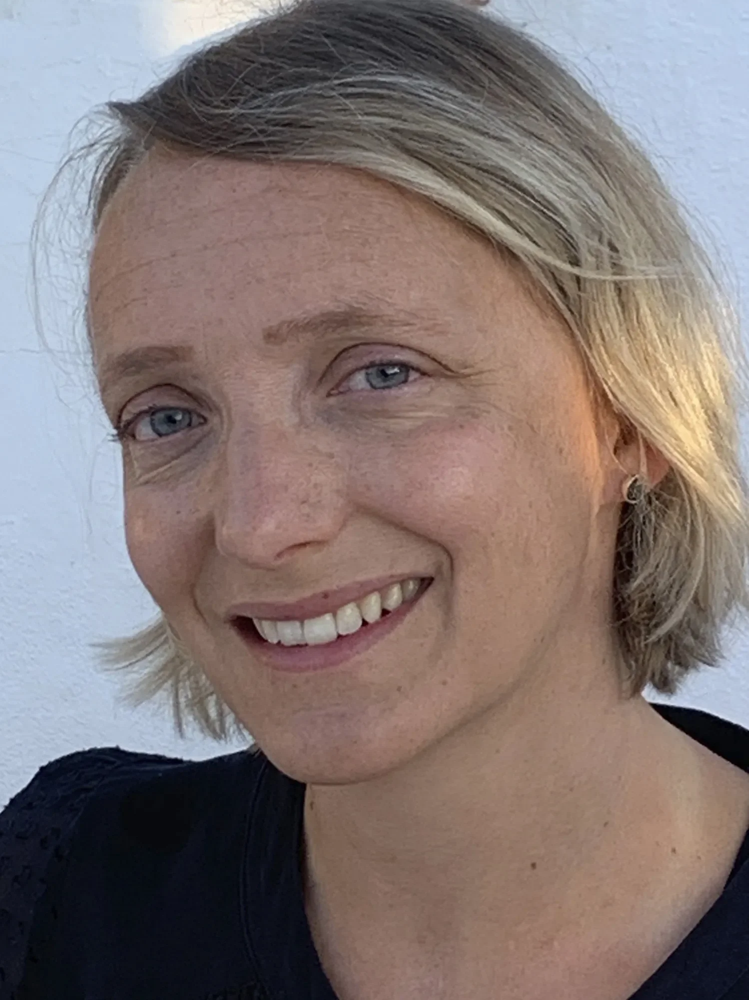 A woman with blonde hair and blue eyes smiling, wearing a black top, against a plain light background.
