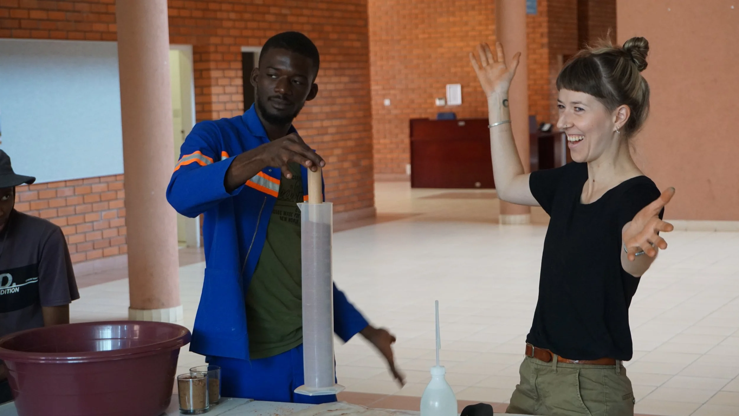 A woman appears excited with her hands raised while a man is looking at a long tube on the table during a science demonstration in an indoor space with brick walls.
