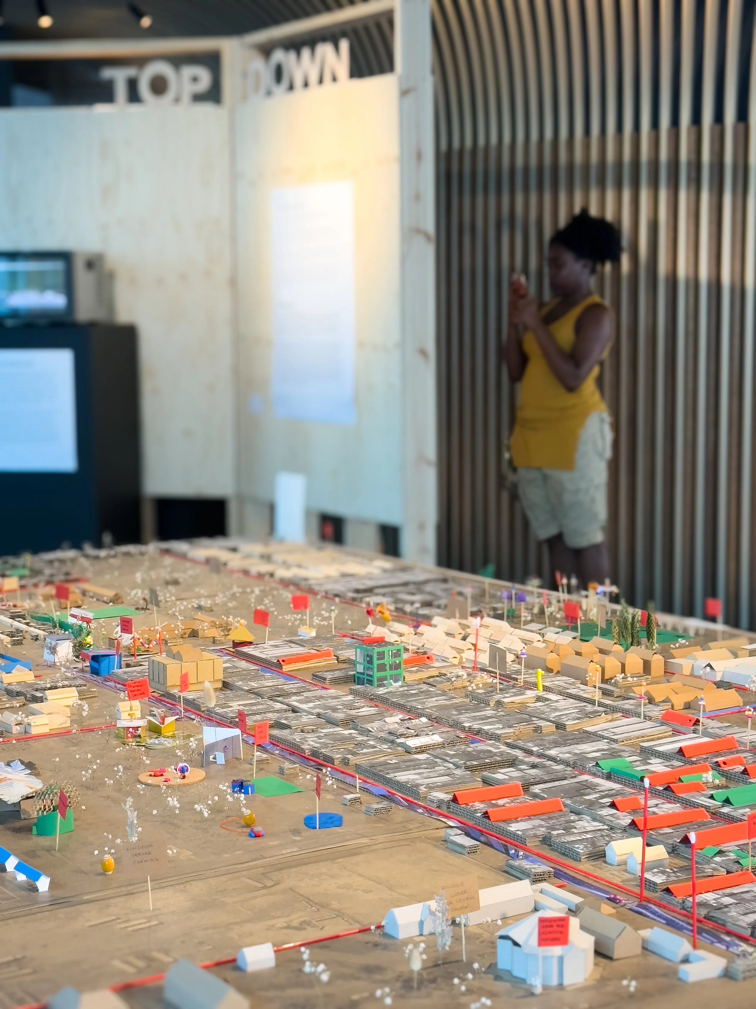A woman stands in front of a detailed scale model of a city layout, with various small buildings, flags, and miniature features, while taking a photo with her phone.