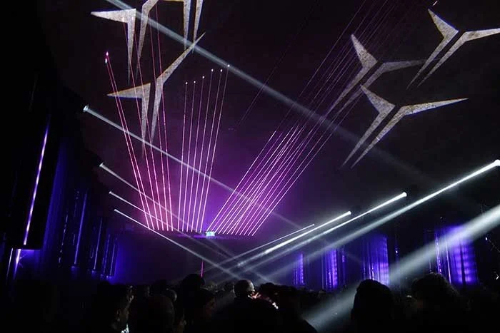 Dark concert or event venue with colorful laser lights and beams, silhouettes of audience members.
