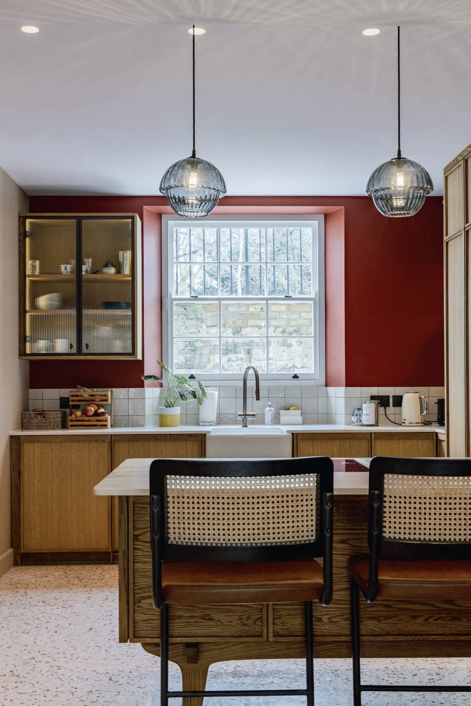 A cozy kitchen with a window above the sink, red accent wall, wooden cabinets, and modern pendant lighting. There are two chairs at a wooden table in the foreground.
