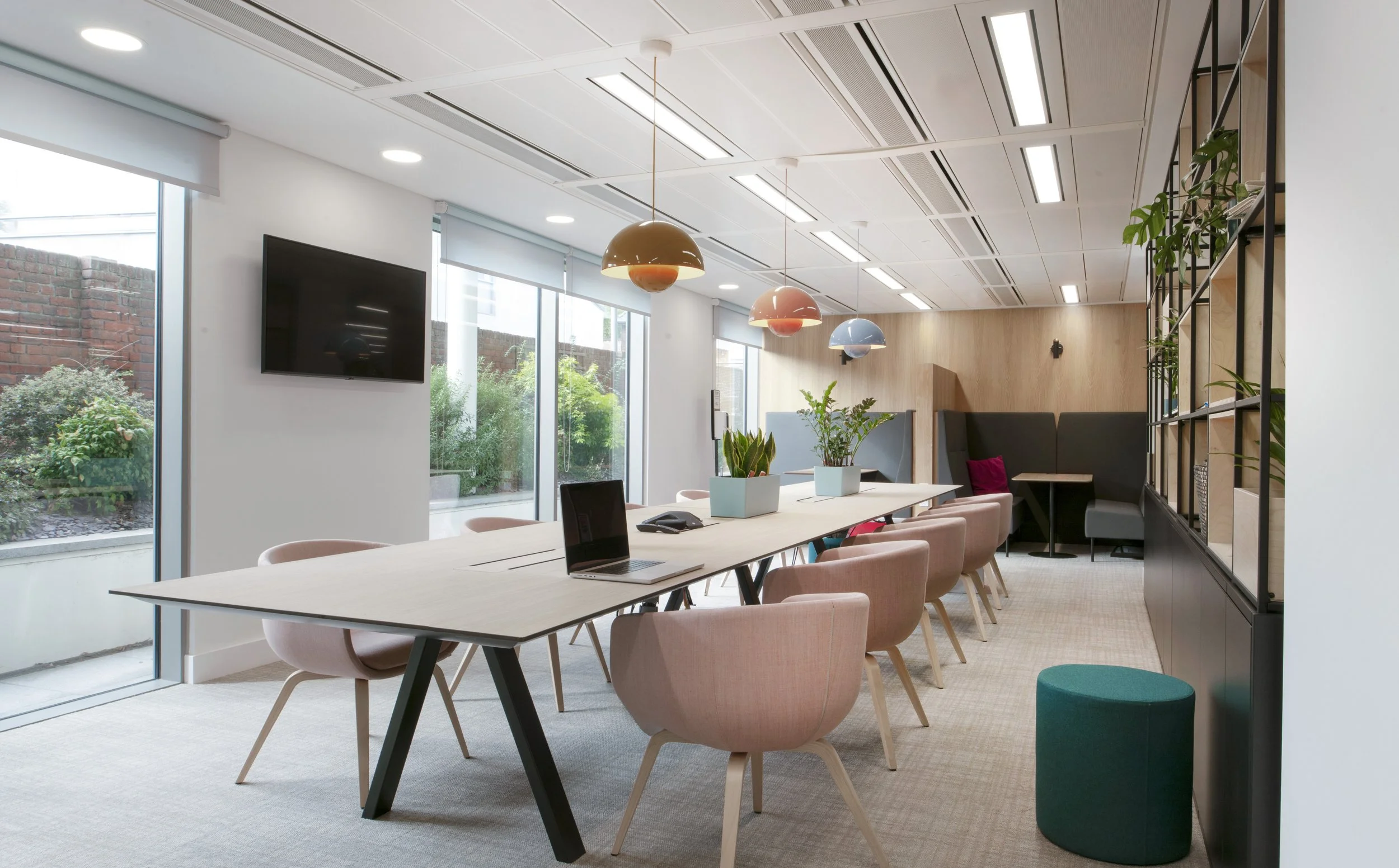 Modern conference room with large table, pink chairs, potted plants, wall-mounted TV, pendant lights, and windows with garden view.