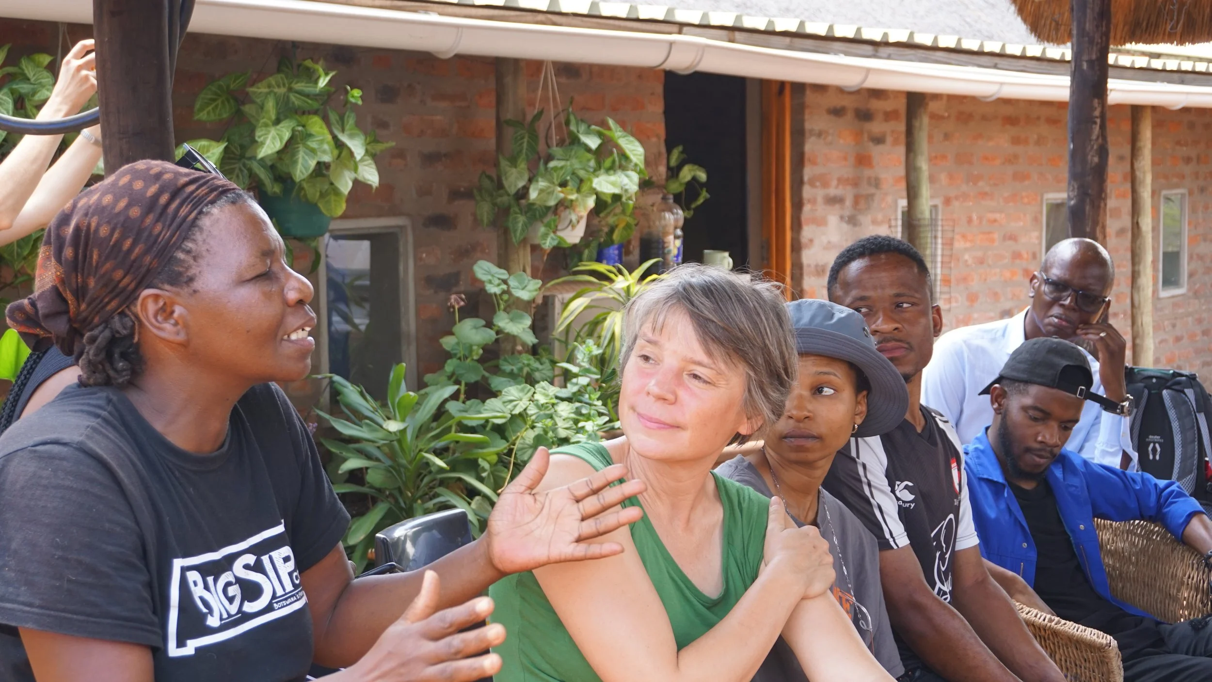 A group of people sitting outdoors, engaged in conversation, with a woman in a black t-shirt and headscarf speaking while others listen, surrounded by green plants and a brick building in the background.