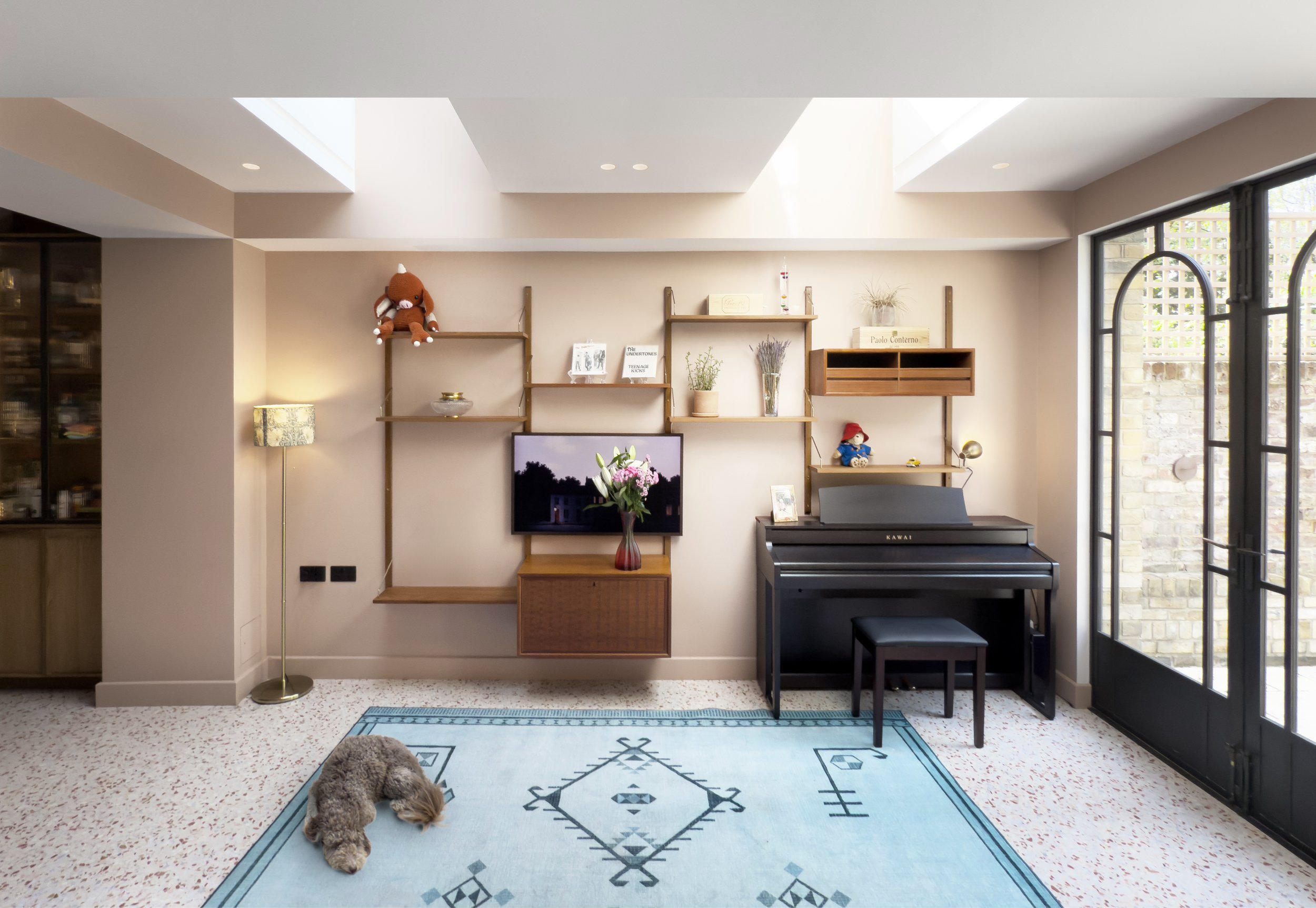 Modern living room with a black digital piano, a small black bench, shelves with decorative items and stuffed toys, a flat-screen TV, a floor lamp, a blue area rug, and a small dog laying on the rug, with large glass door windows on the right.