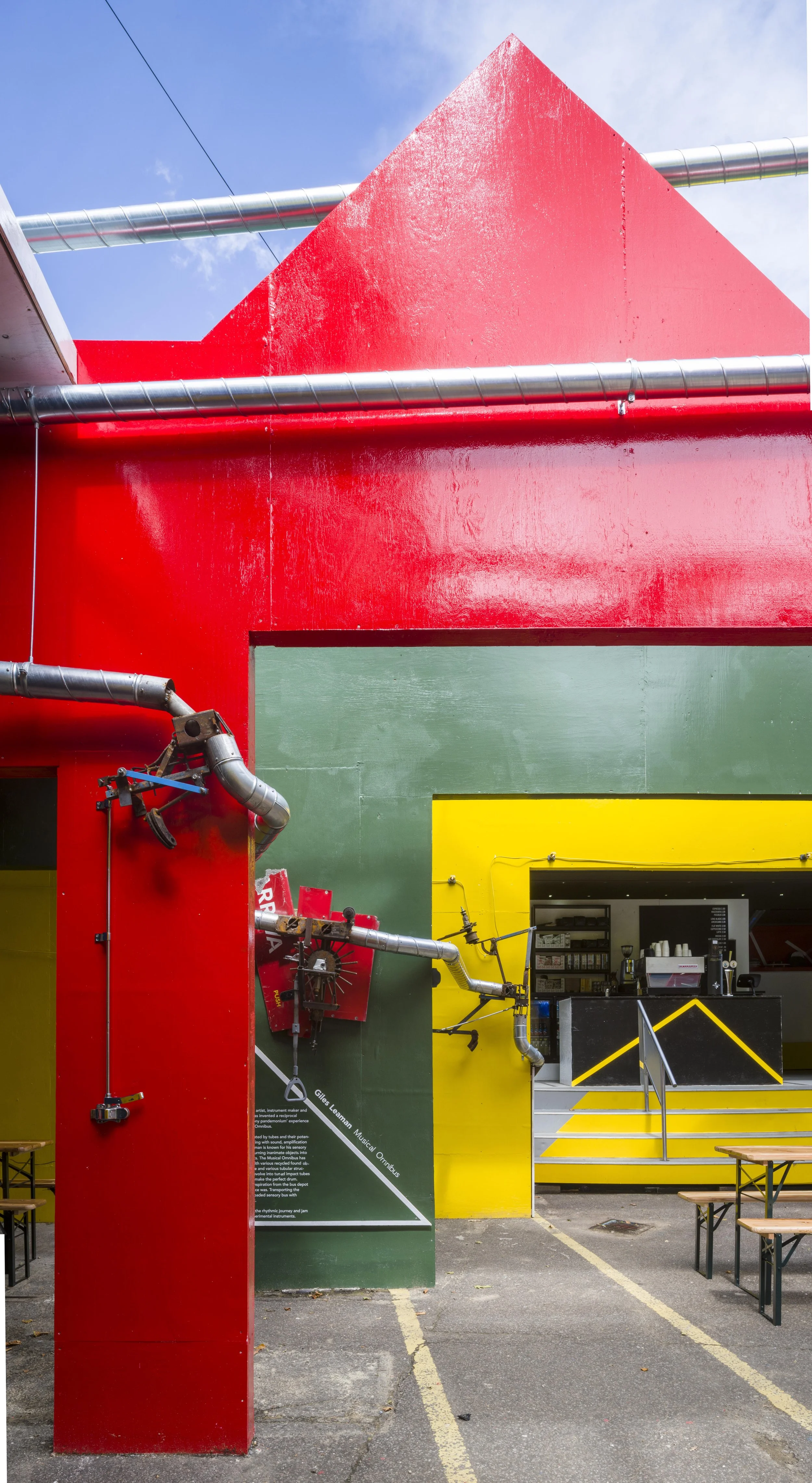 Colorful building with red, green, yellow walls and pipes, with a small outdoor seating area and a food stand.