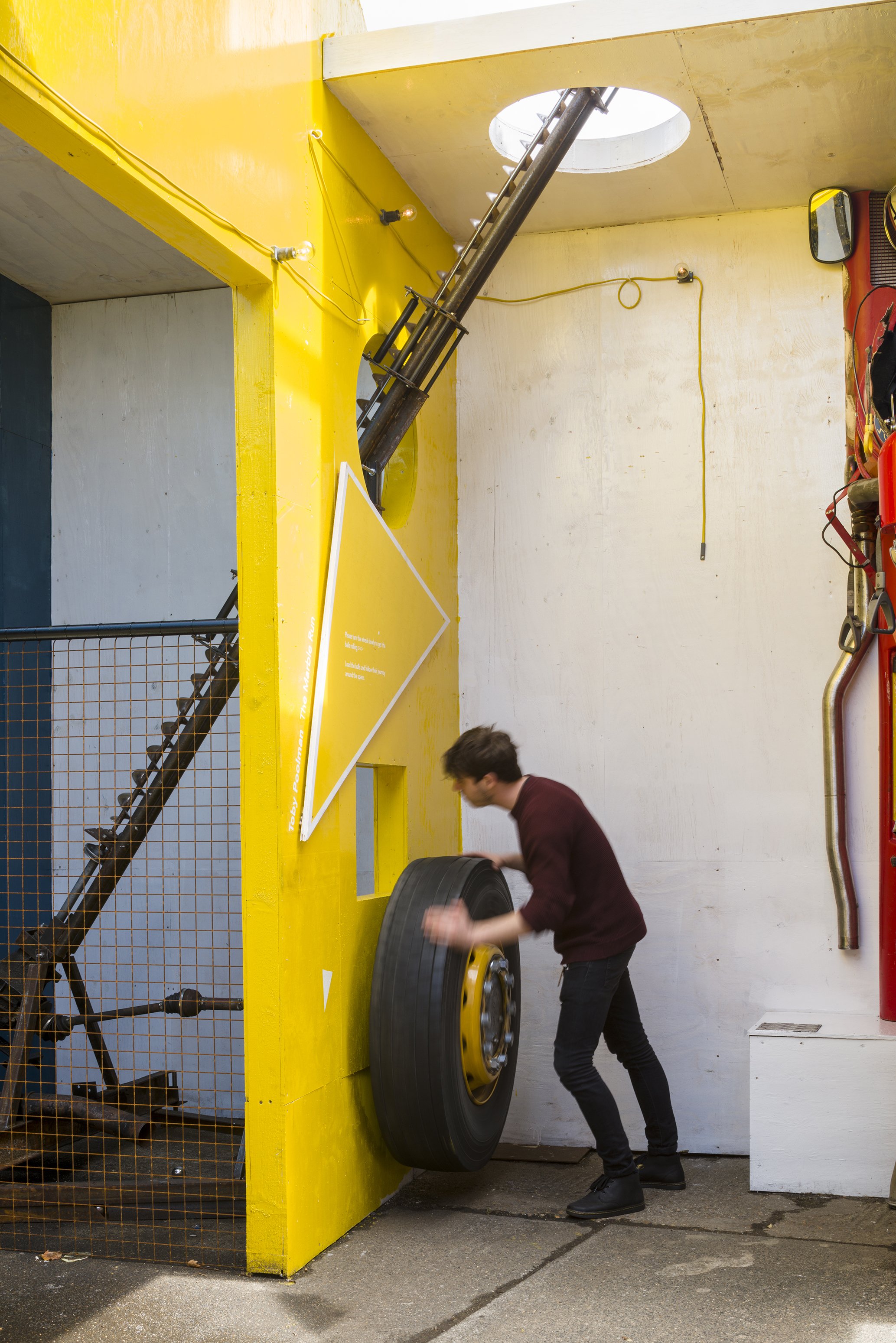 A person in a maroon sweater and black pants is holding a large motorcycle tire in front of a yellow wall with an opening.