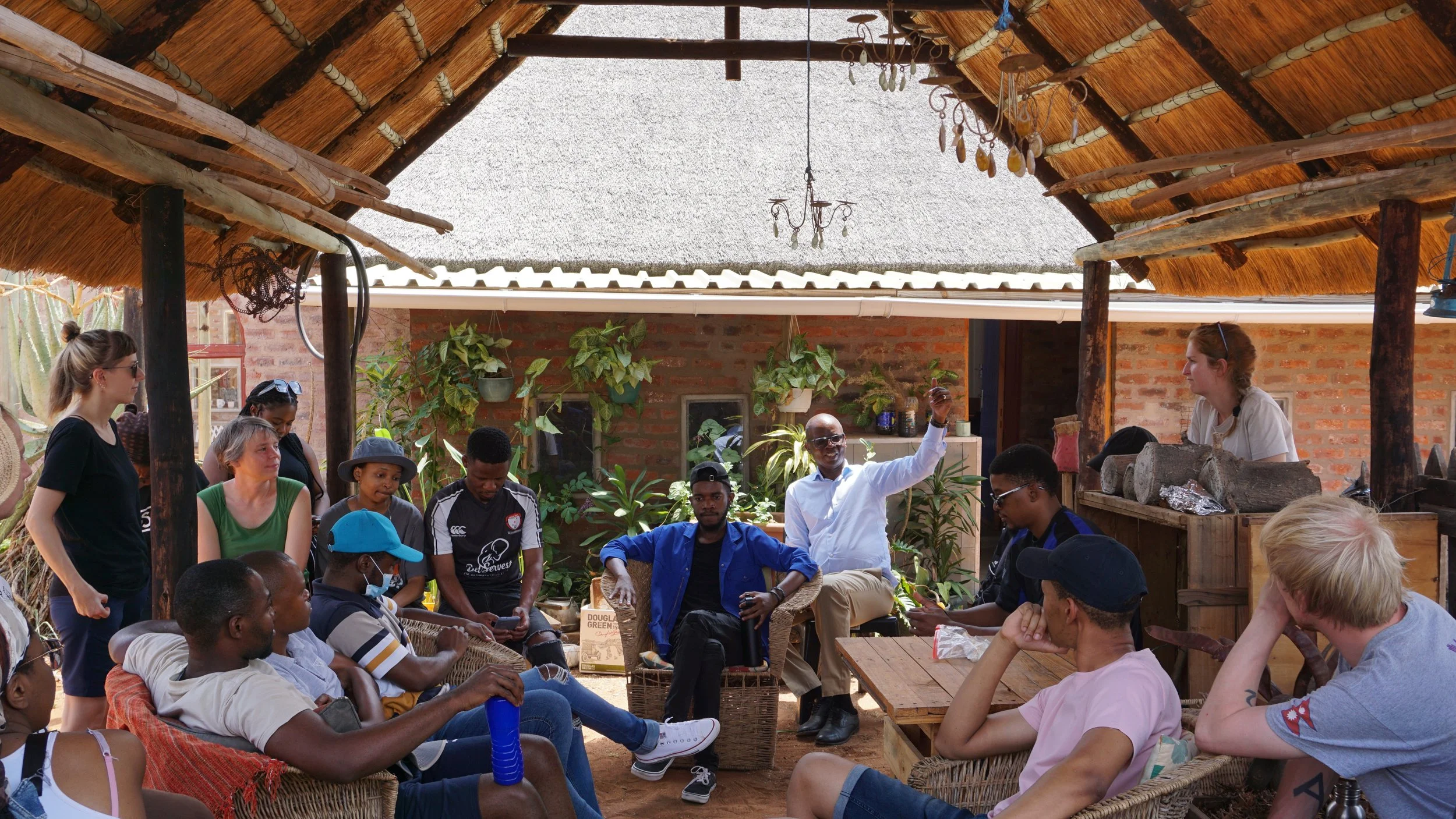Group of diverse people gathered under a thatched roof outdoor space, listening to a man in a white shirt talking and gesturing, with everyone seated on chairs and sofas, surrounded by plants and rustic decor.