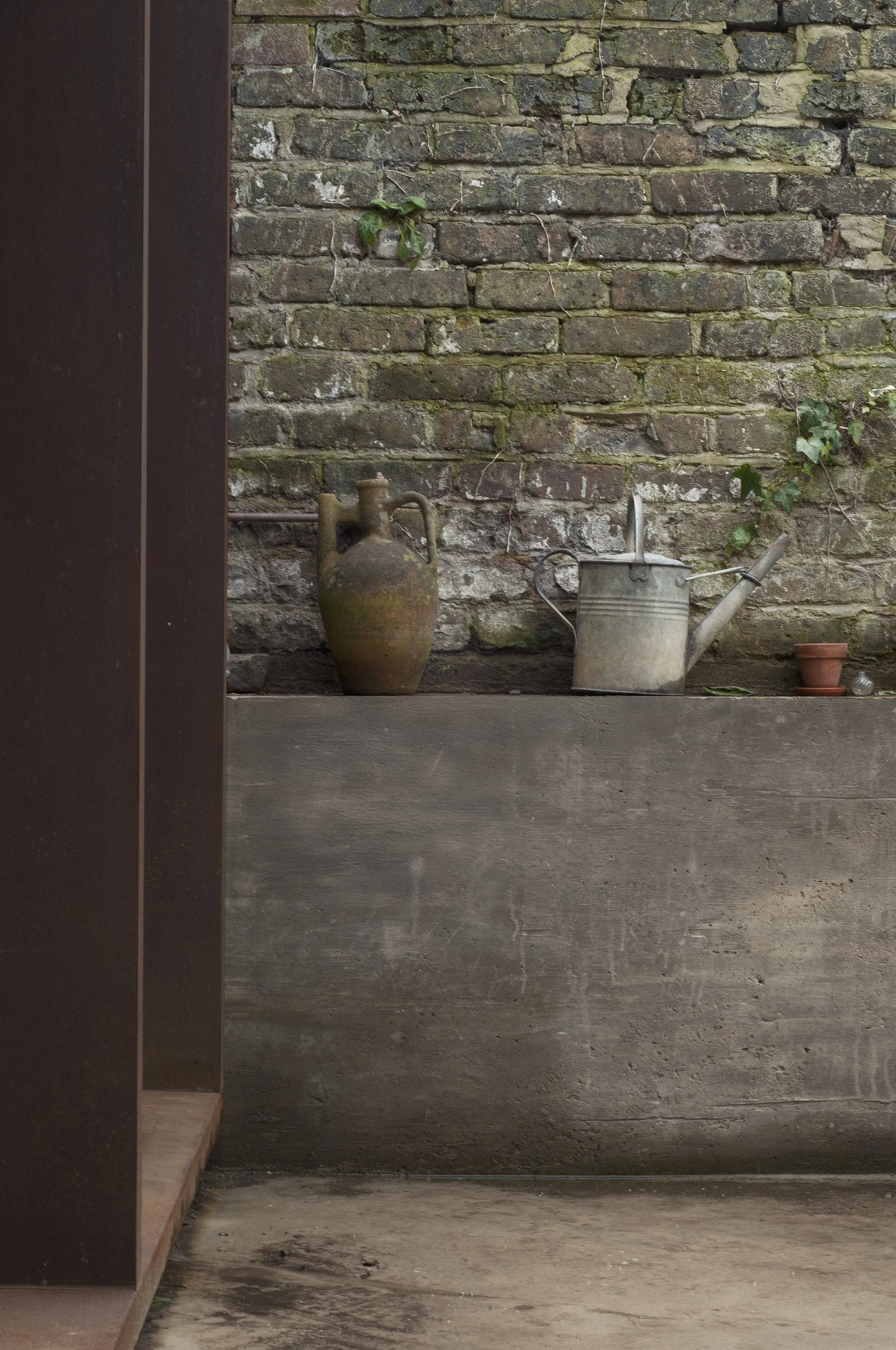 A rustic scene with an old clay jug, a metal watering can, a small terracotta pot, and some green plants against a weathered brick wall.