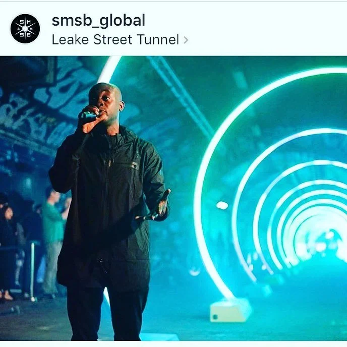 A person holding a microphone standing in front of an illuminated circular structure at Leake Street Tunnel.