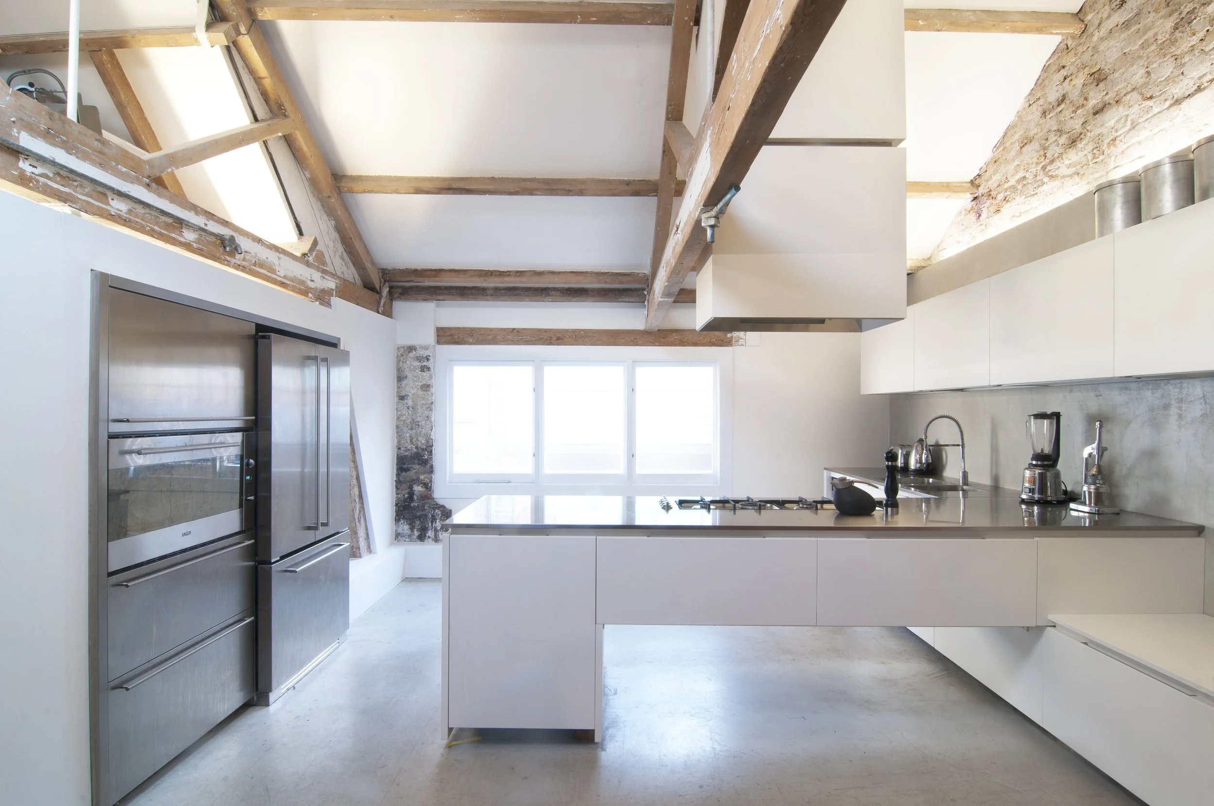 Modern kitchen with stainless steel refrigerator, white cabinets, and a concrete backsplash, featuring a large window, an island, and exposed wooden beams on the ceiling.