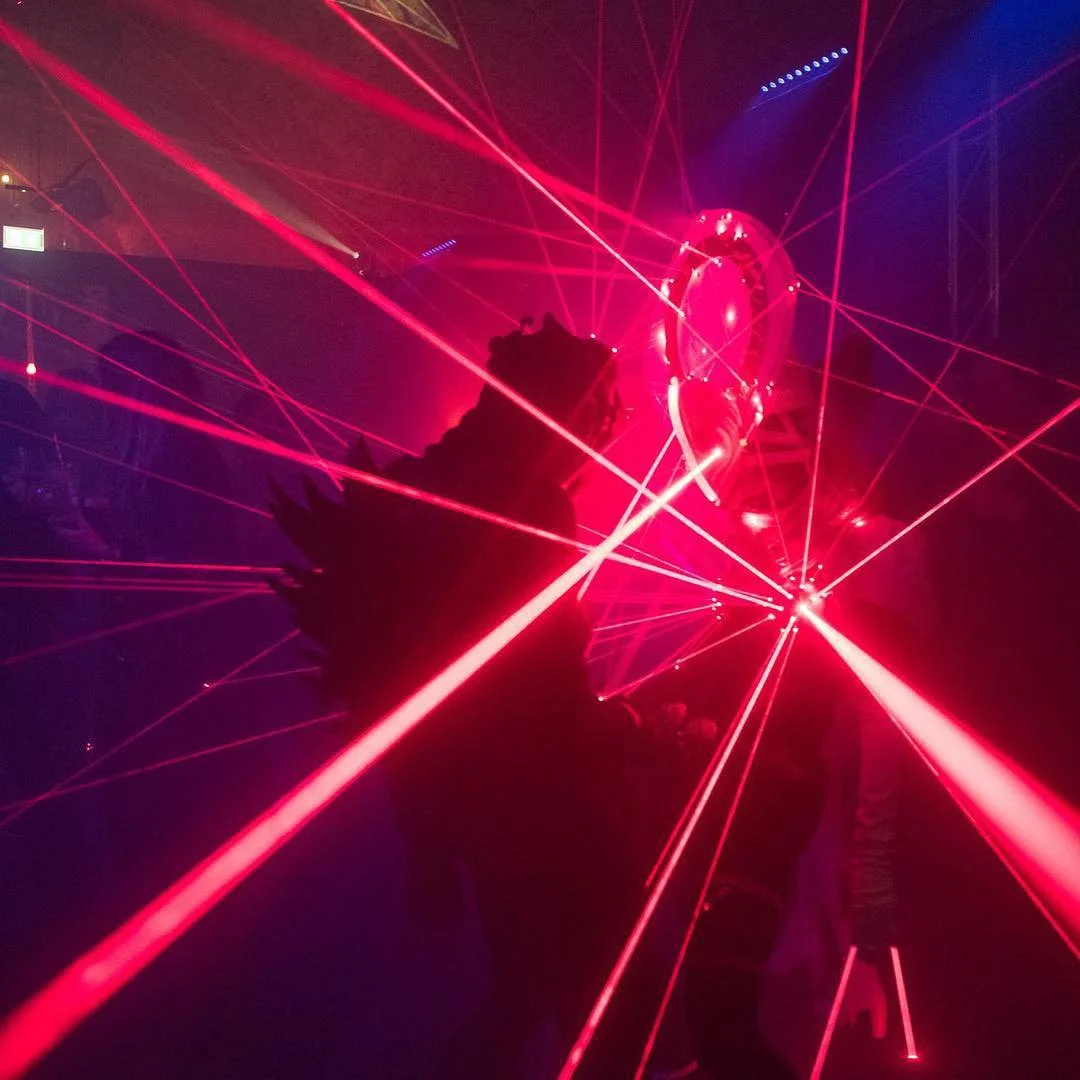 A person at a laser tag or entertainment venue surrounded by red laser beams and colorful lighting, with a large, illuminated object in the background.
