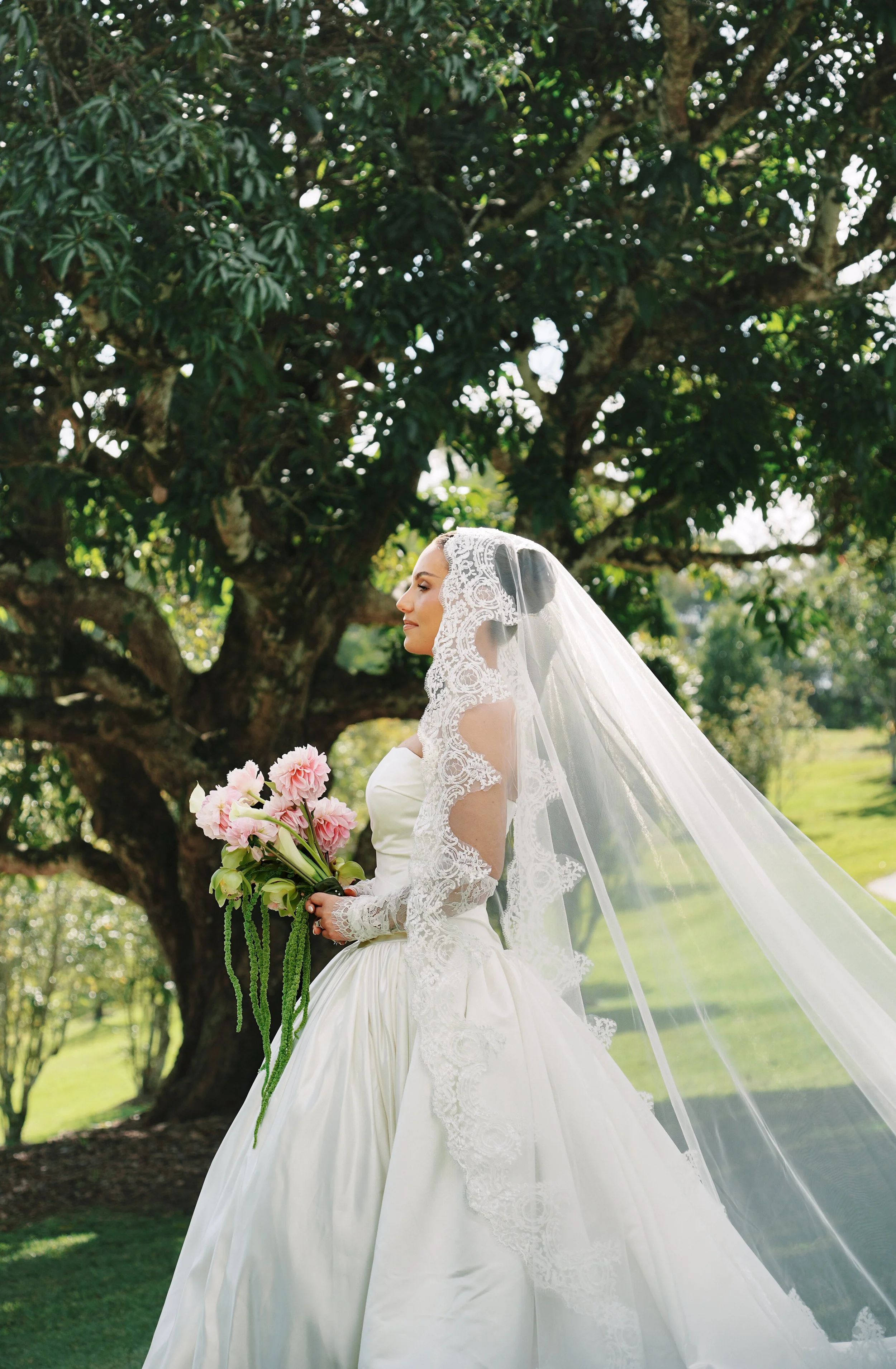 Bride in a white wedding gown with lace sleeves and a veil holding a bouquet of pink flowers outdoors under a large leafy tree.