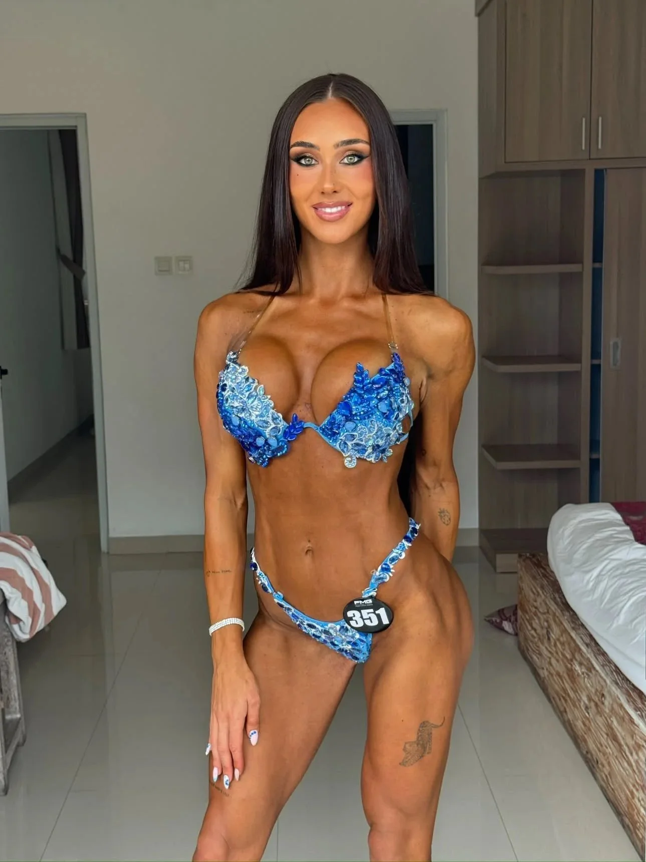 Woman with long dark hair and makeup standing indoors, wearing a blue bikini with embellishments, a medal with the number 351, and showing tattoos on her left arm and thigh, in a room with a bed and wooden furniture.