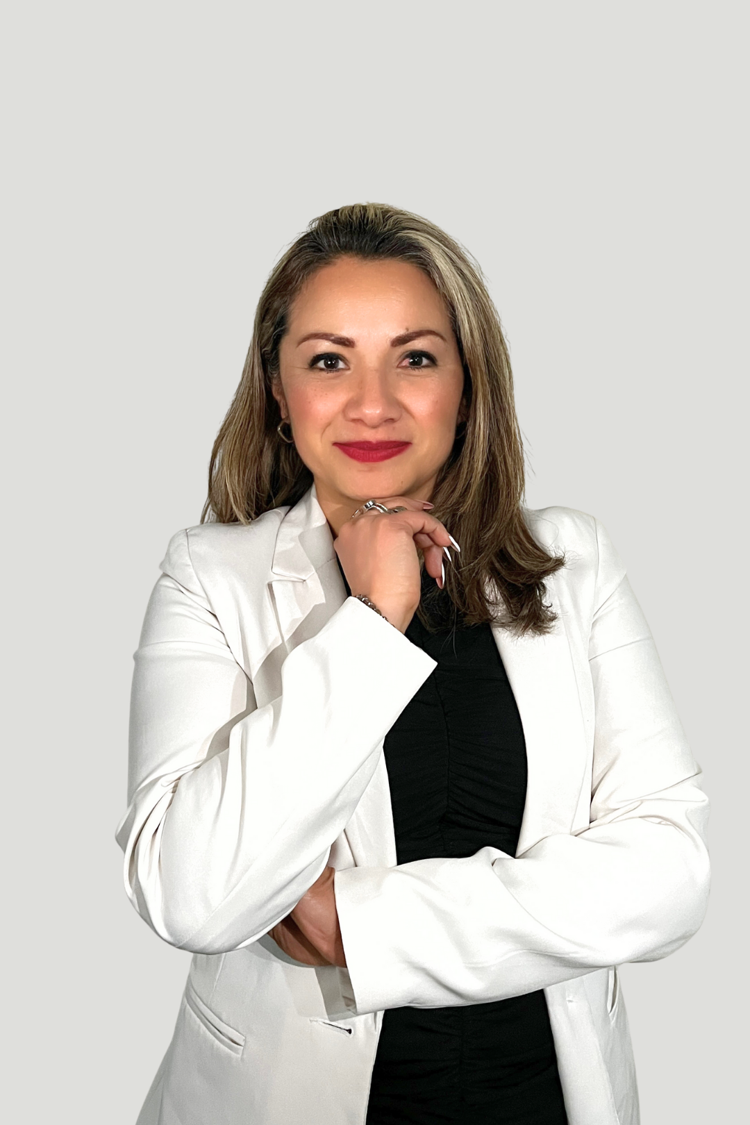 Mujer con cabello castaño claro y labios rojos, vestida con chaqueta blanca y blusa negra, posando con la mano en la barbilla, frente a fondo gris.