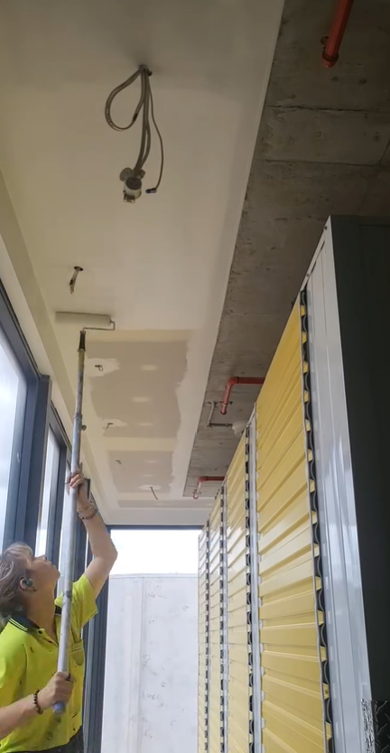 A woman in a yellow shirt using a telescoping pole to reach the ceiling near the light fixture in a room with storage lockers and large windows.