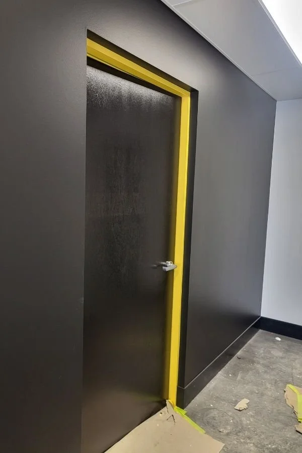 Black door with yellow protective tape framing the edge, located in a modern gray walled interior with a dark floor.