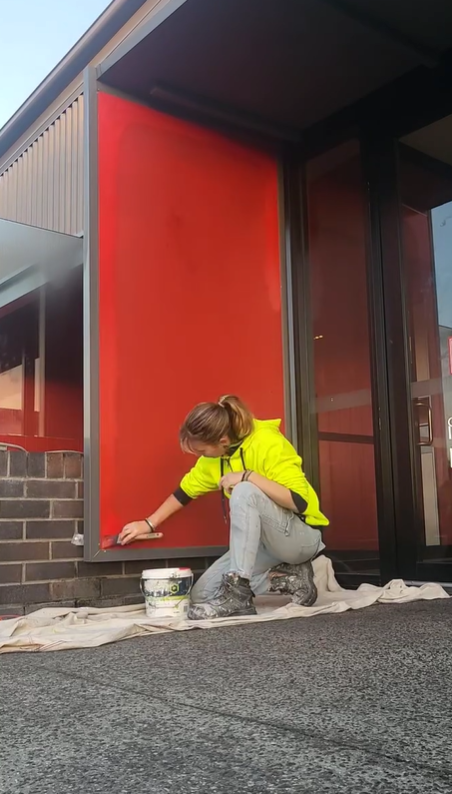A woman in a yellow jacket is painting a section of a building red near the entrance, crouched down with painting supplies on the ground.
