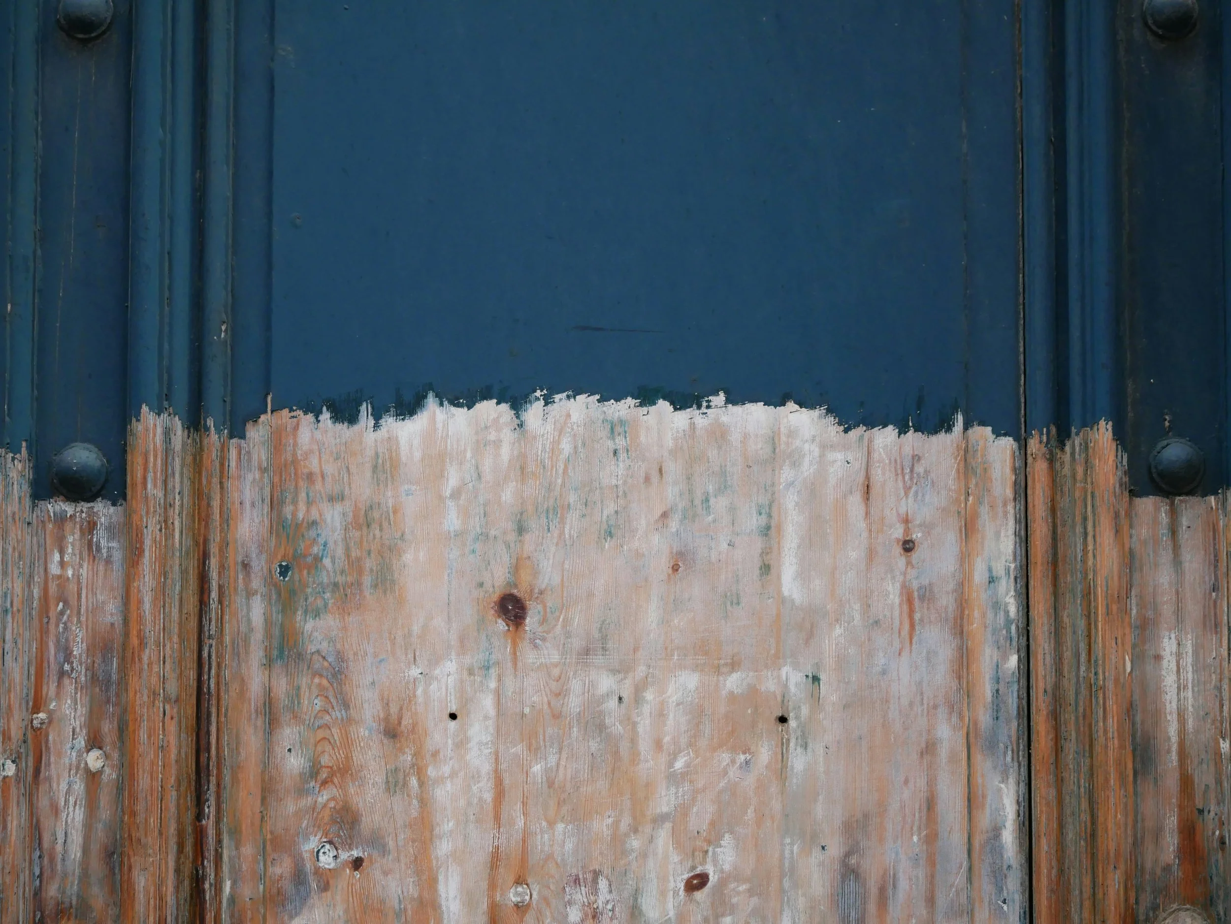Close-up image of a weathered wooden door with a painted blue upper section and a lower section with exposed, peeling wood.