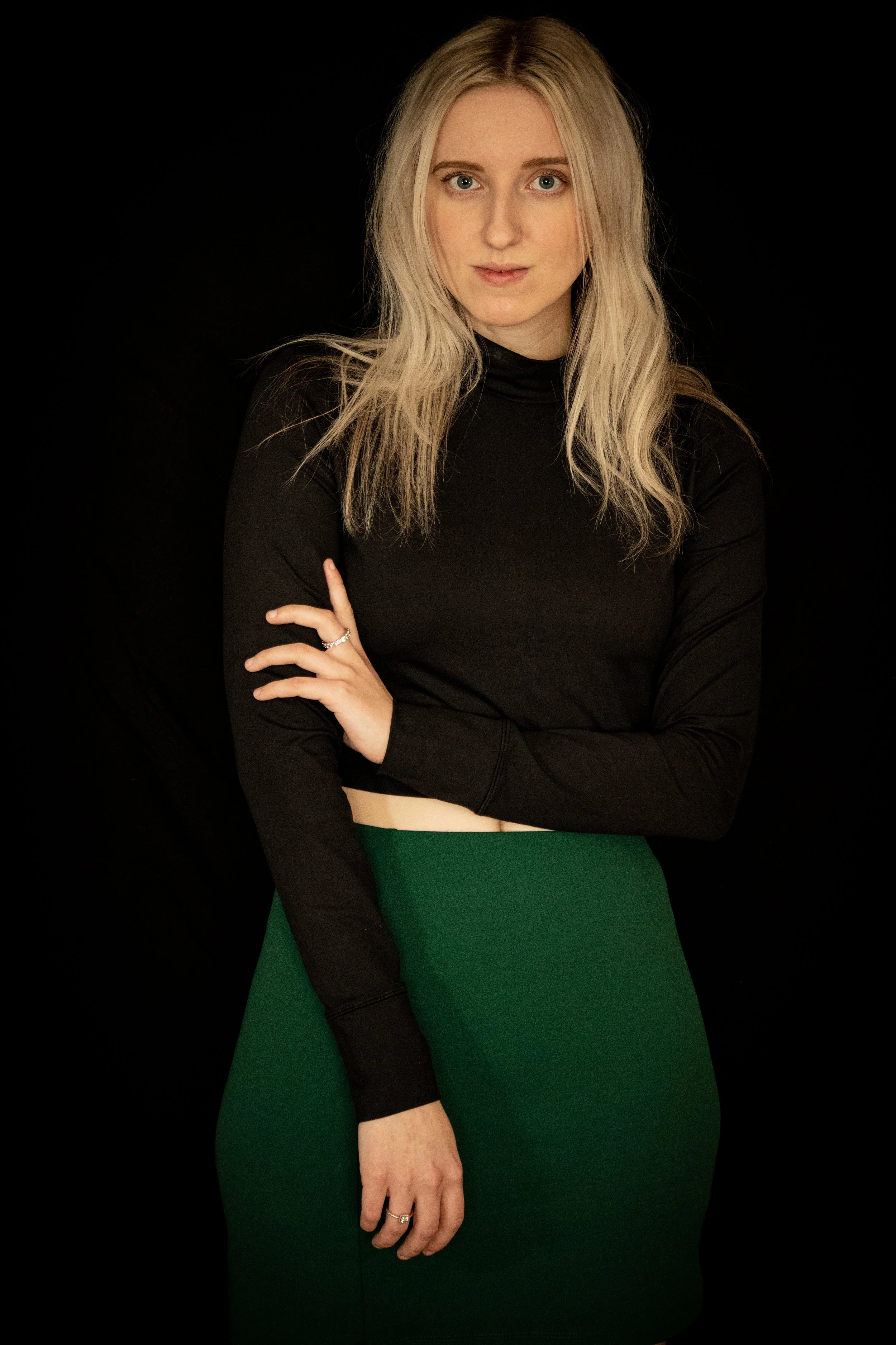 A young woman with long blonde hair wearing a black long-sleeve top and a green skirt, posing against a black background.