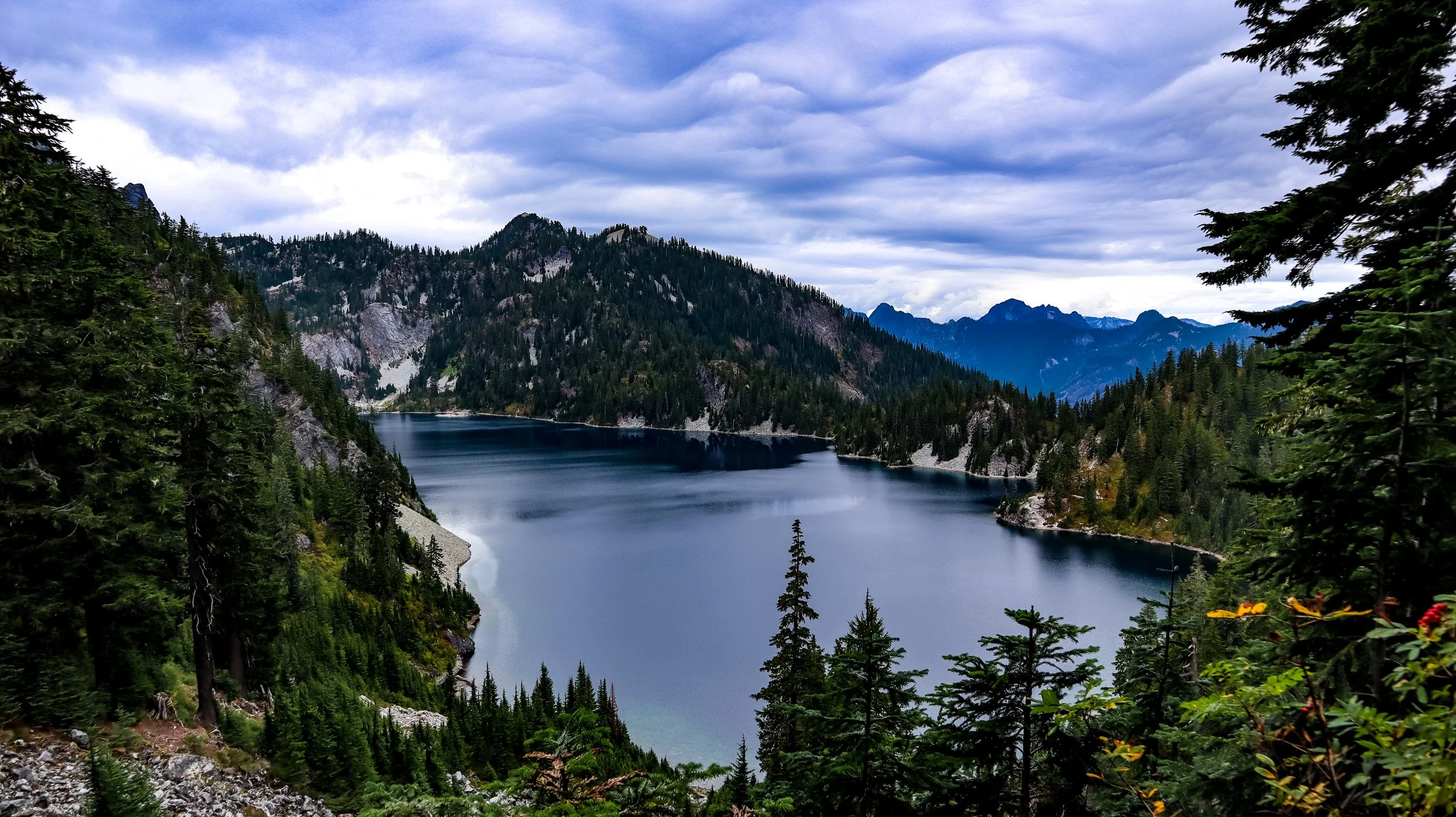 A scenic view of a lake surrounded by dense evergreen forests and mountain ranges under a cloudy sky.