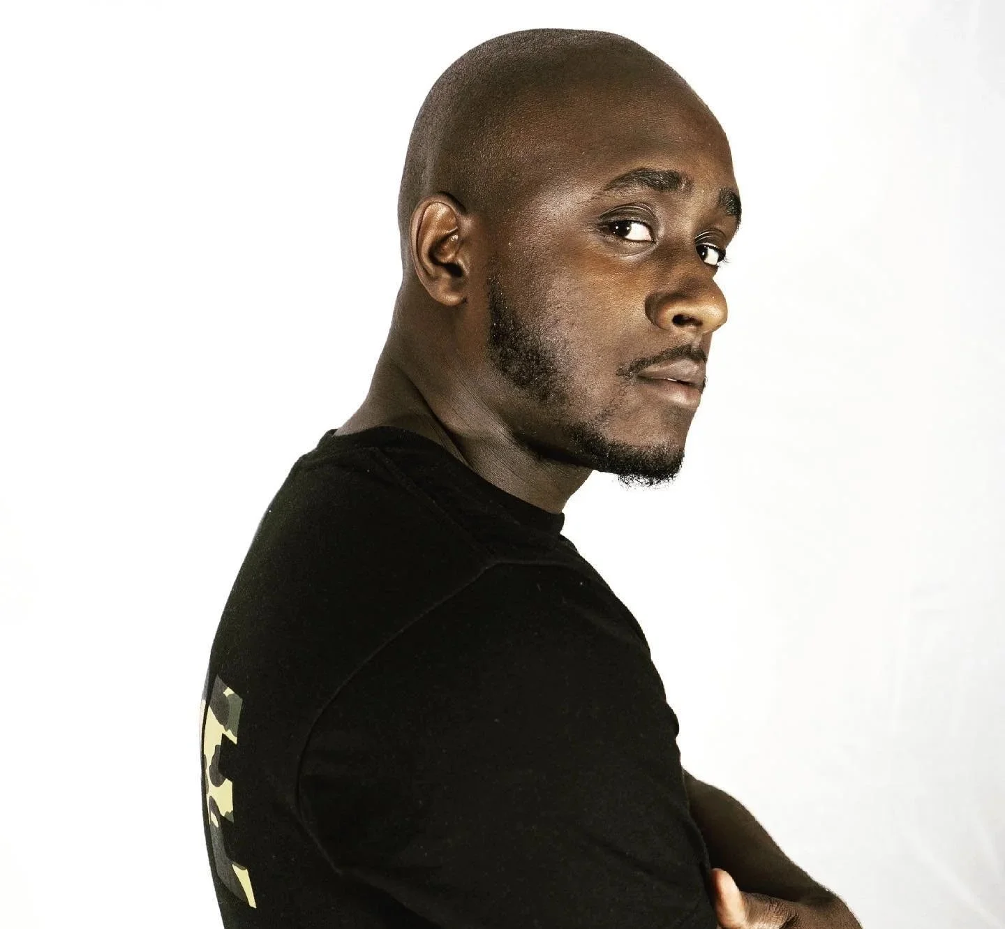 A man with a dark complexion, shaved head, and beard, wearing a black T-shirt, standing against a white background, looking over his shoulder.
