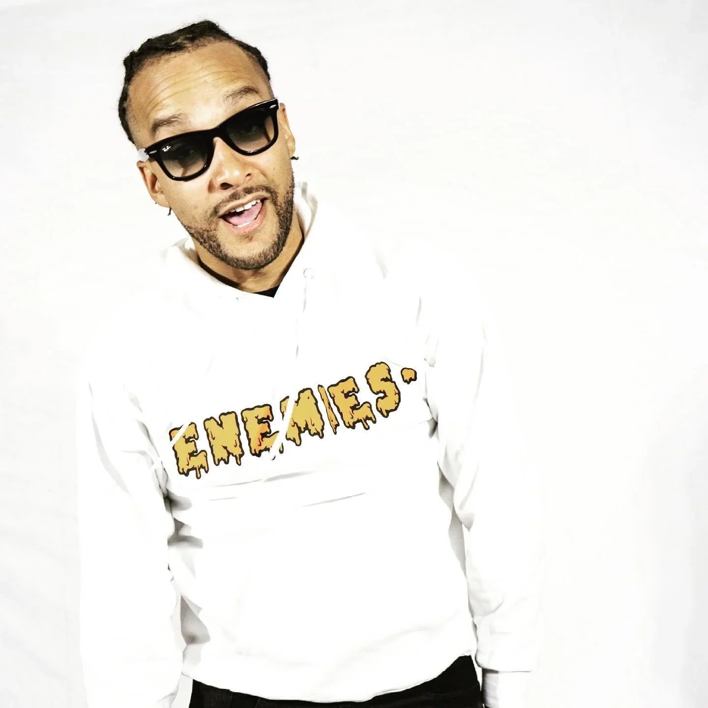 A man wearing sunglasses and a white hoodie with the word 'ENEMIES' in stylized yellow and brown letters, standing against a white background.