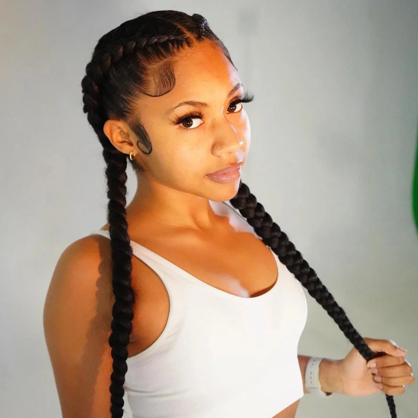 A young woman with braided hair, wearing a white sleeveless top, looking at the camera, holding her braid with her left hand, against a plain background.