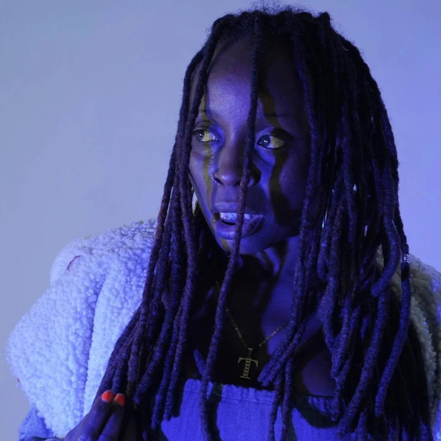 A woman with long dreadlocks, wearing a white textured jacket and a necklace with a letter T pendant, is illuminated by blue and purple lighting.