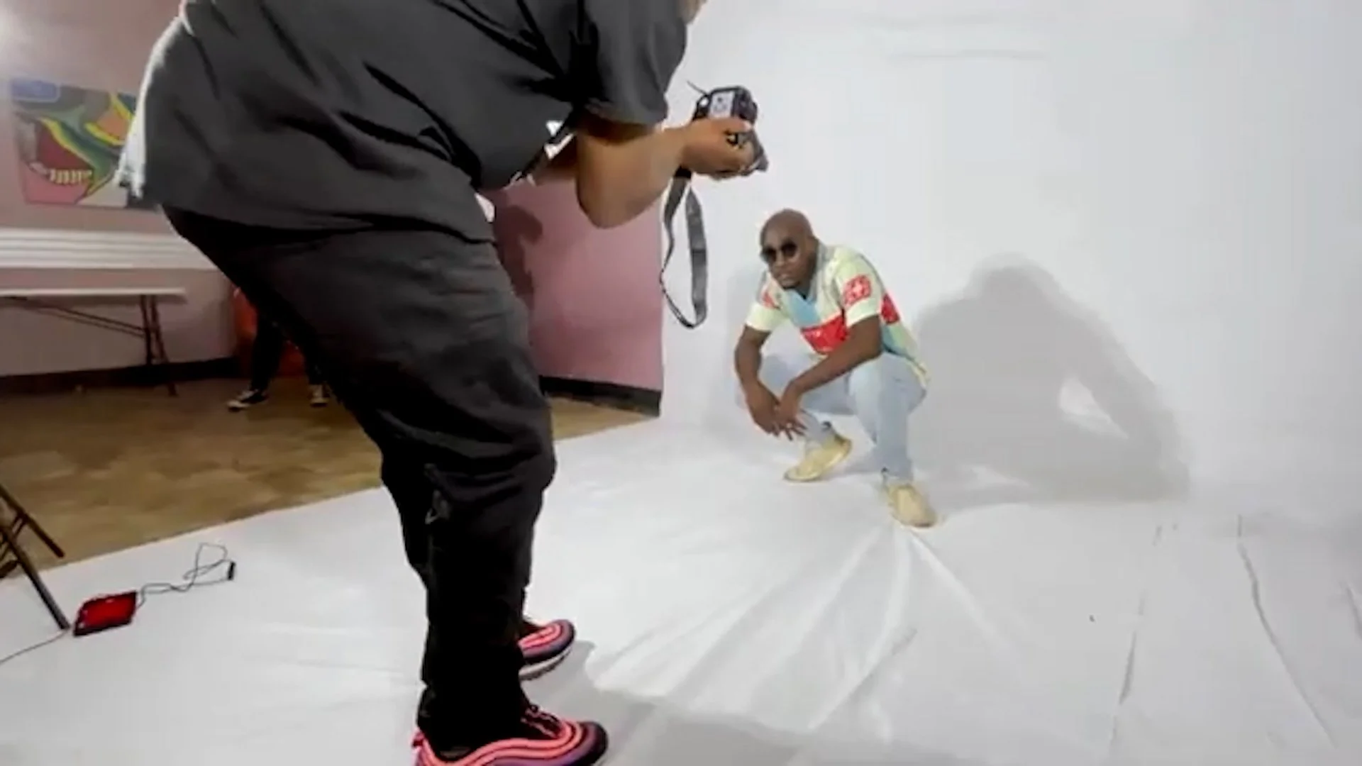 Photographer taking a picture of a man squatting with sunglasses in front of a white backdrop inside a room.