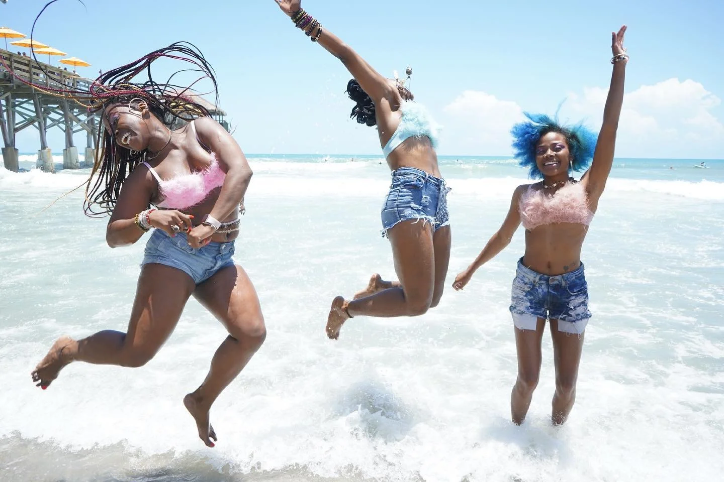 Three women joyfully jumping in the ocean waves at the beach, with a pier in the background, under a bright blue sky.