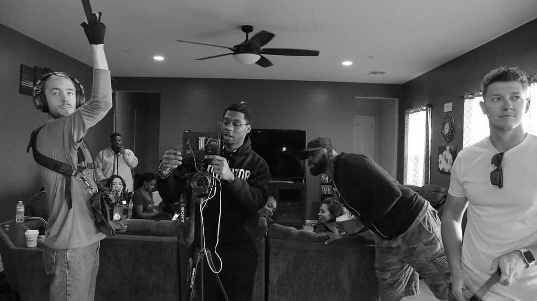 A group of people on a film or photo shoot in a living room, with one man adjusting equipment, others engaging in tasks, and some seated on a couch.