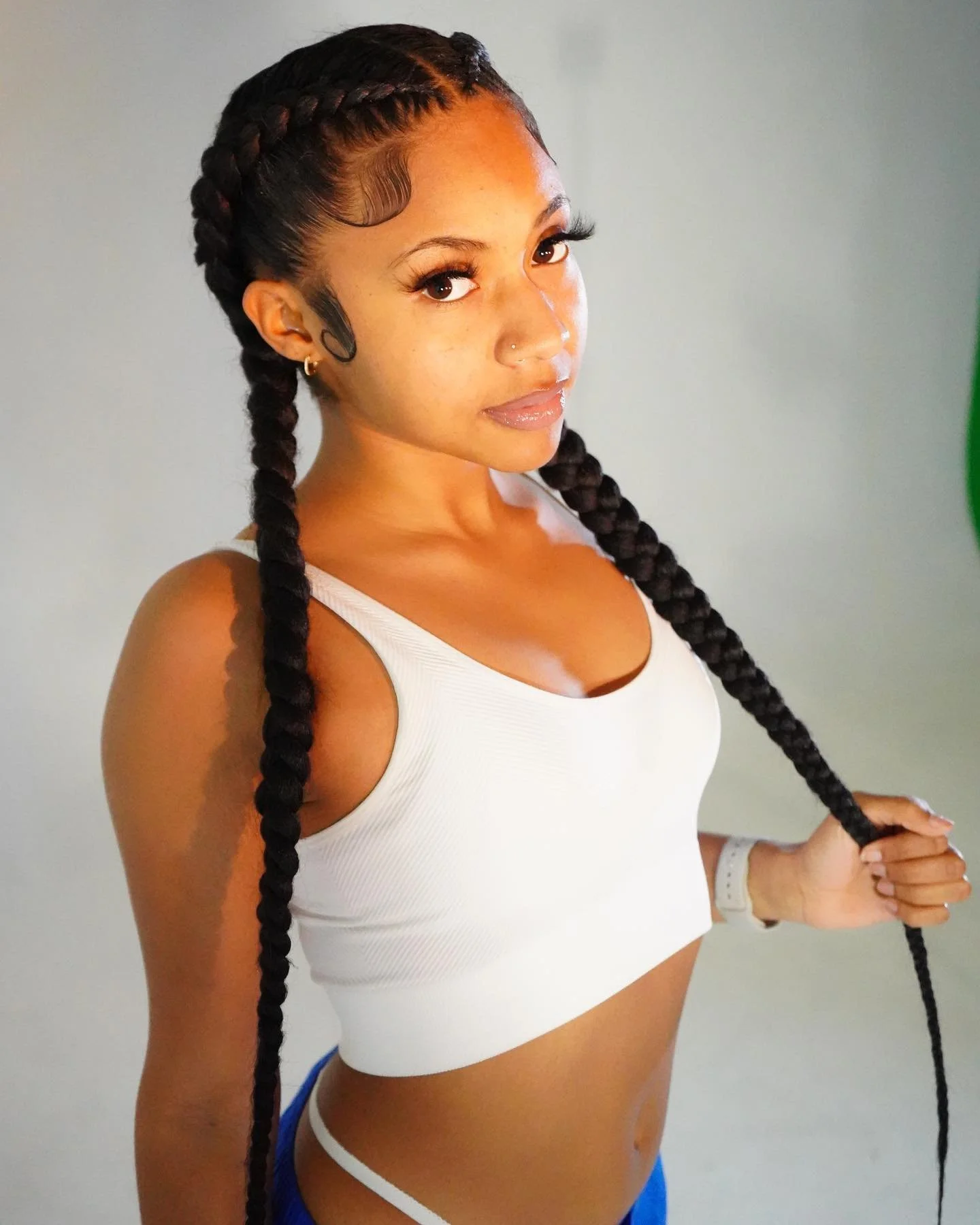 A young woman with long braided hair, wearing a white sports bra and blue shorts, standing against a plain background.