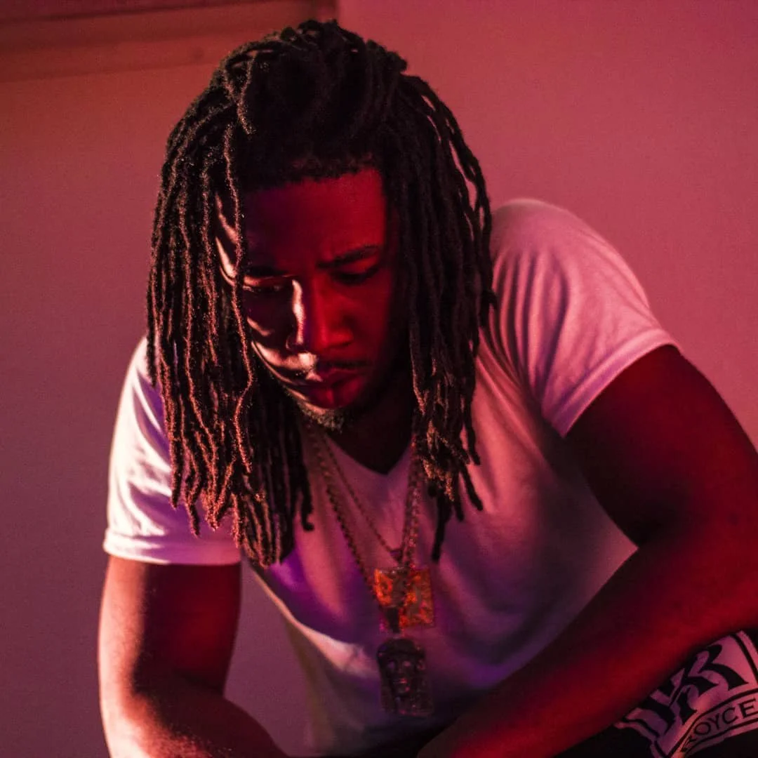 Young man with long dreadlocks and jewelry, wearing a white T-shirt, looking down against a pinkish background.