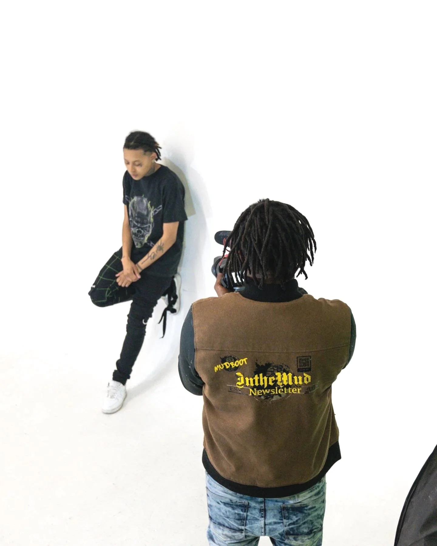 A person wearing a brown vest with yellow text taking a photo of a young man with dreadlocks against a plain white background.