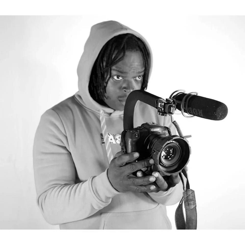 A person with dreadlocks and wearing a hoodie, holding a Canon camera with a microphone attached, looking focused.