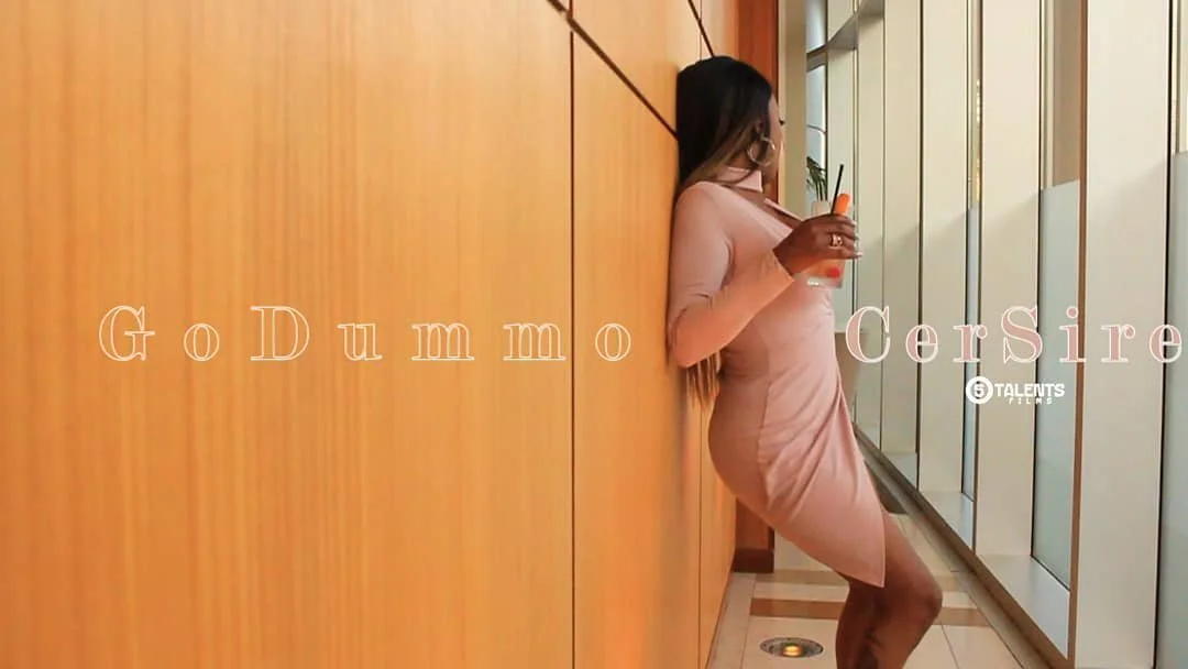 A woman in a beige dress leaning against a wooden wall, drinking from a glass with a straw, next to large windows.