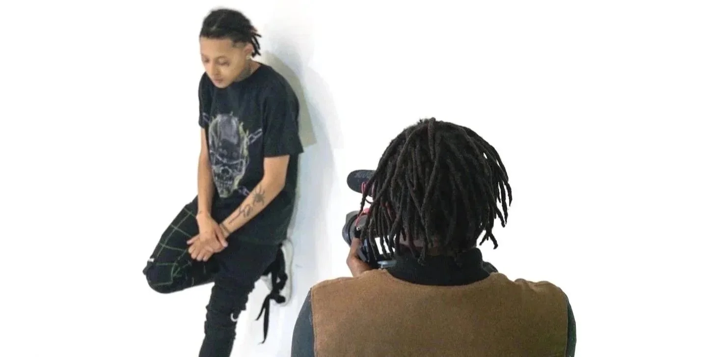 A young man sitting on a stool facing a person with dreadlocks using a camera, against a plain white background.