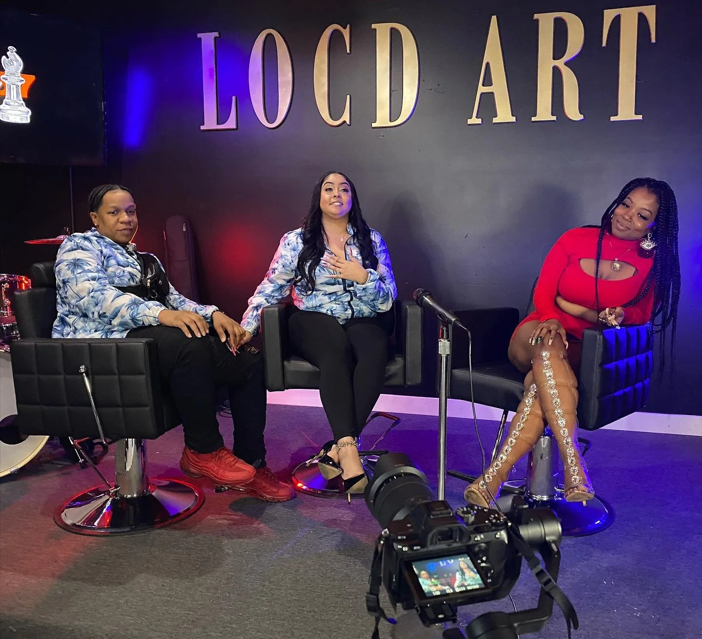 Three women sitting on black chairs during a talk show at LOC'D ART. The woman in the middle has long black hair, wearing black pants and high heels, and has her hand on her chest. The woman on the right has long braided hair, wearing a red dress with transparent lace-up shoes, and is smiling. The woman on the left is wearing a blue patterned jacket and black pants, holding the hand of the woman in the middle. There is a camera in front of them, recording the scene.