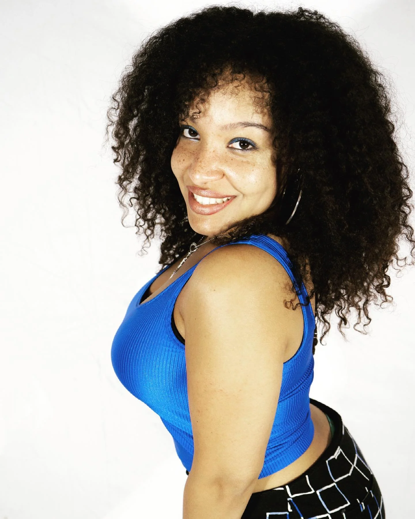 A woman with curly hair wearing a blue sleeveless top and smiling at the camera against a white background.