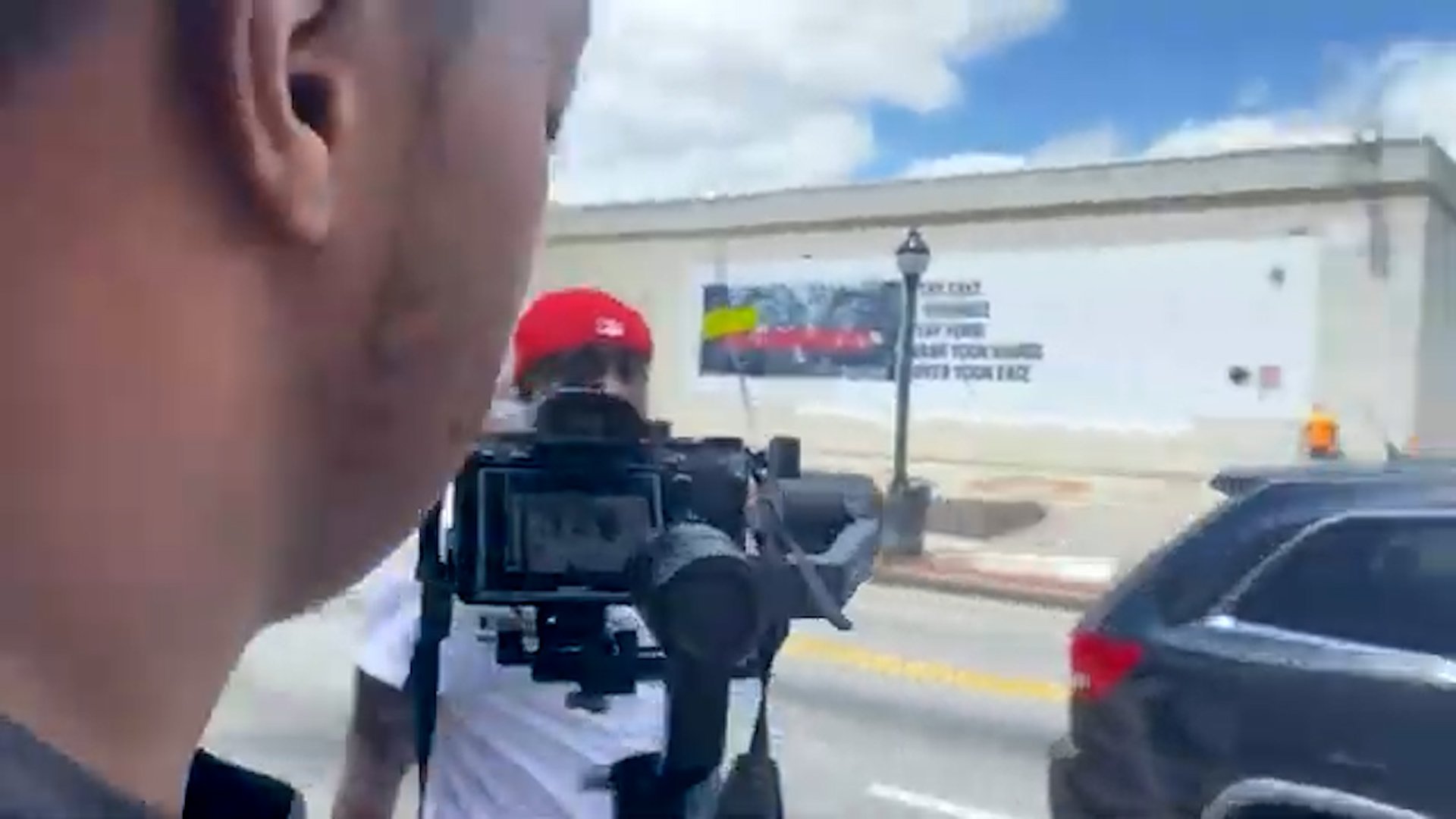 Close-up of a person's face on the left side of the frame, with a street scene in the background including a person in a red hat, a camera person, cars on the road, and a white building with a billboard.