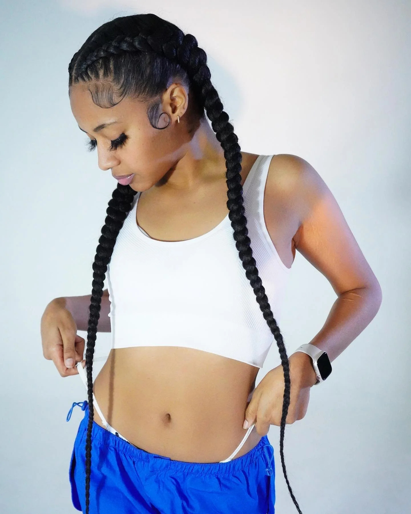 Woman with long braided hair wearing a white sports bra, blue shorts, an Apple Watch, and looking down.