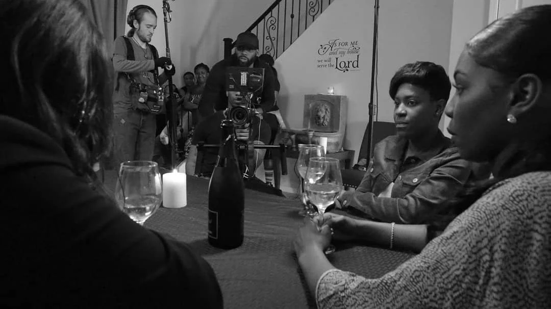 A scene from a filming set showing three women at a table with wine glasses, a candle, and a bottle. Behind them, crew members operate cameras and equipment, with a staircase and wall decor in the background.