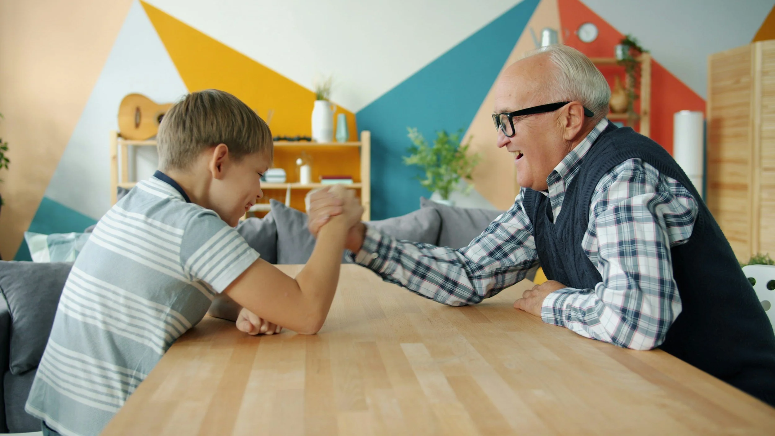 Young boy and elderly man engaged in arm wrestling at a dining table, smiling and enjoying each other's company in a colorful living room.