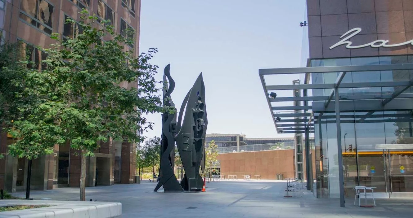 Modern urban plaza with a large abstract black sculpture in the center, surrounded by trees, glass buildings, and benches, on a clear day.