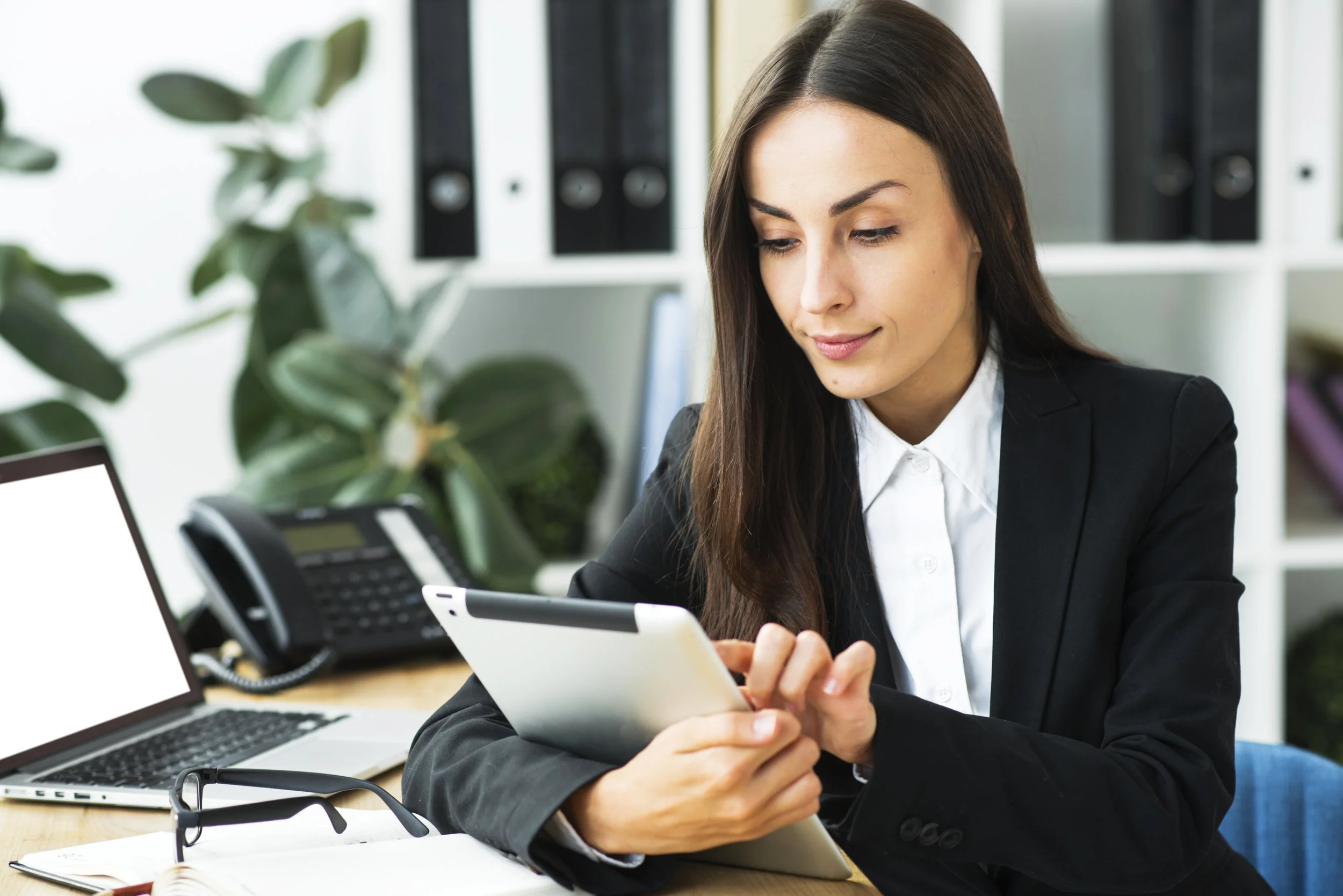 A woman in a black blazer sitting at an office desk, looking at a tablet, with a laptop, landline phone, glasses, and notebook nearby, in a professional office setting.