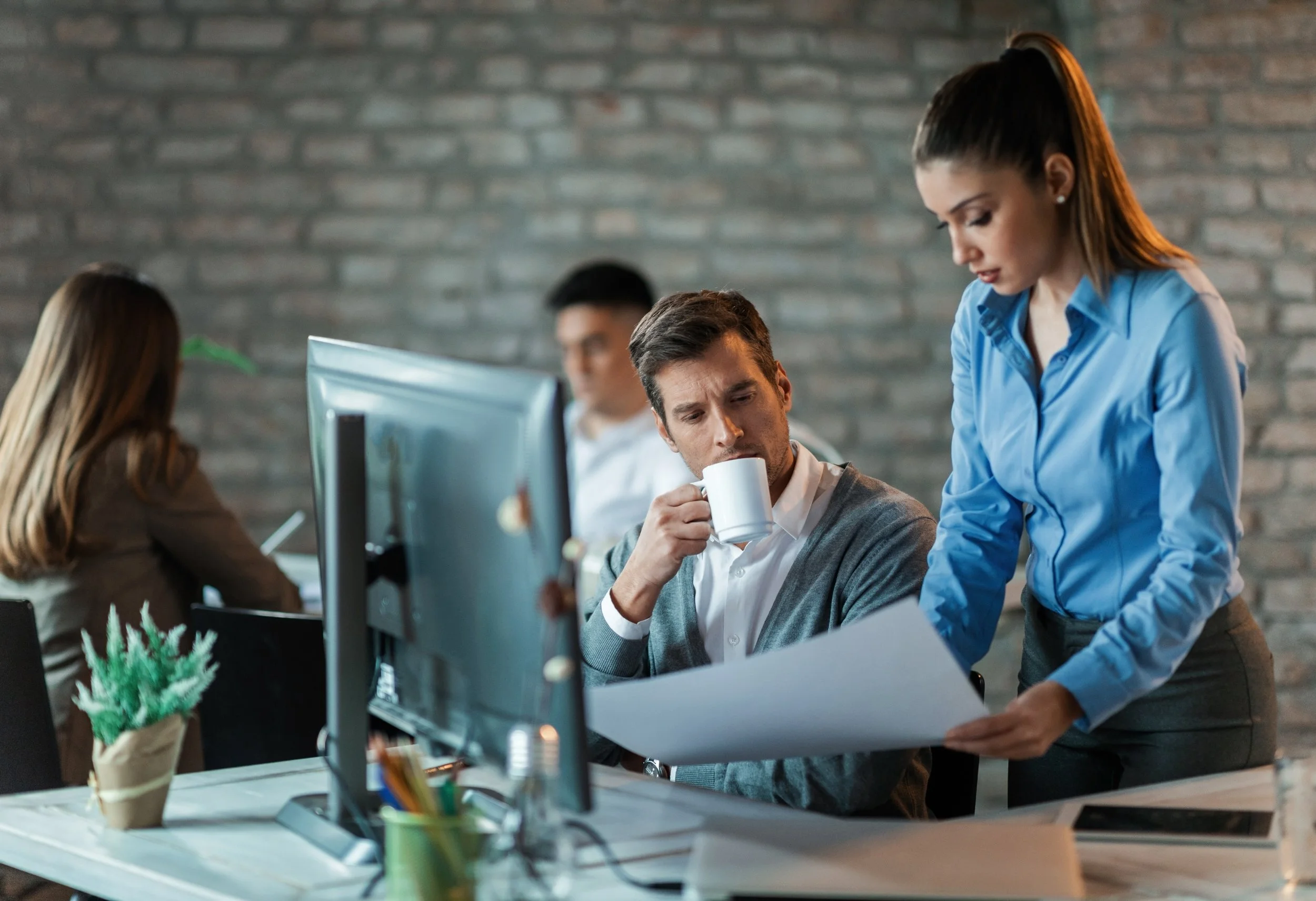 Businesswoman explains paperwork to man drinking coffee in office.