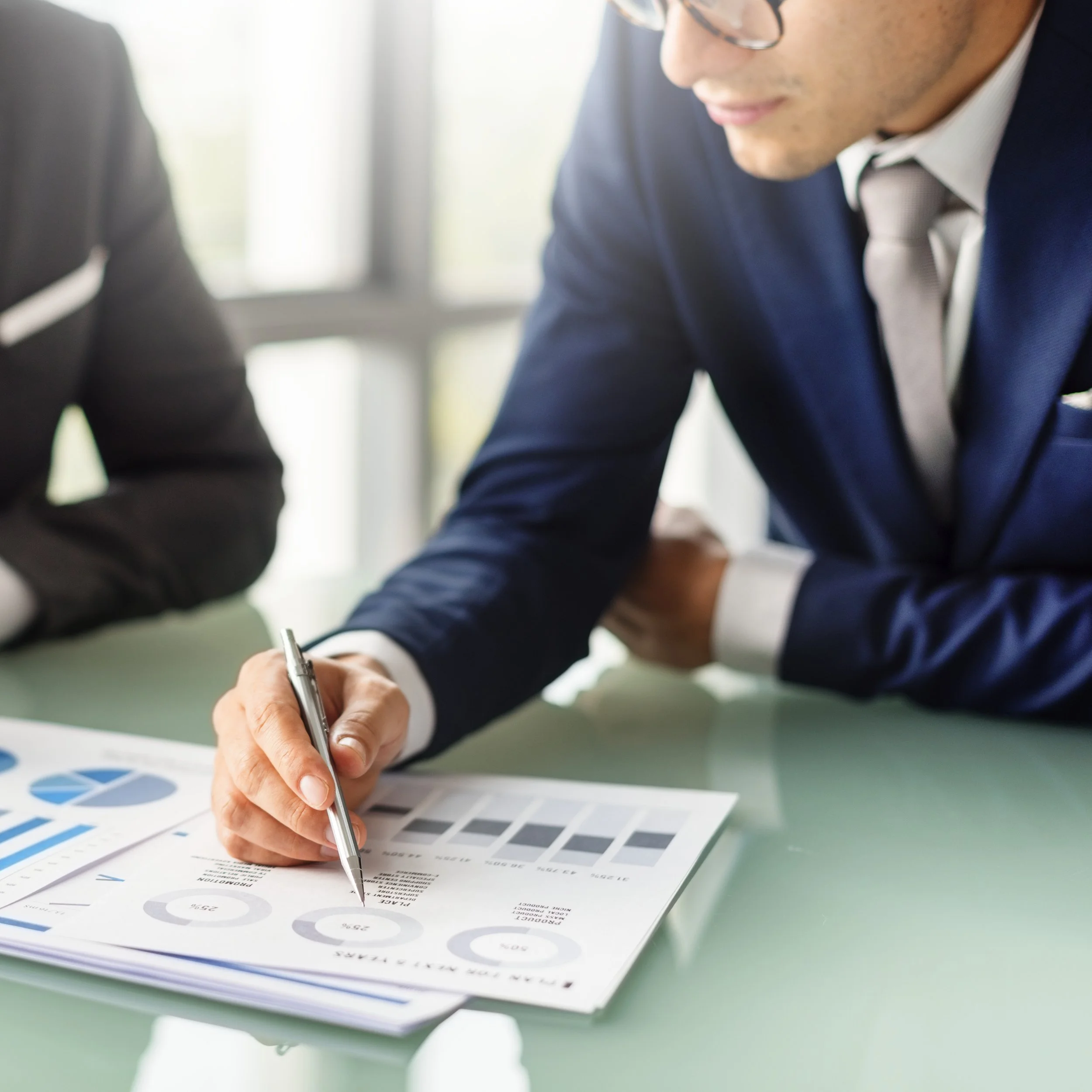 Business professional in a suit pointing at printed data charts and graphs on a table during a meeting or presentation.