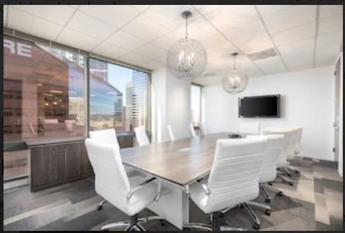 Modern conference room with a large wooden table, eight white office chairs, large windows, and a wall-mounted TV.