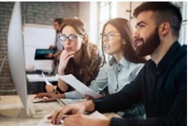 Three young professionals working together in an office, looking at a computer screen with focused expressions.