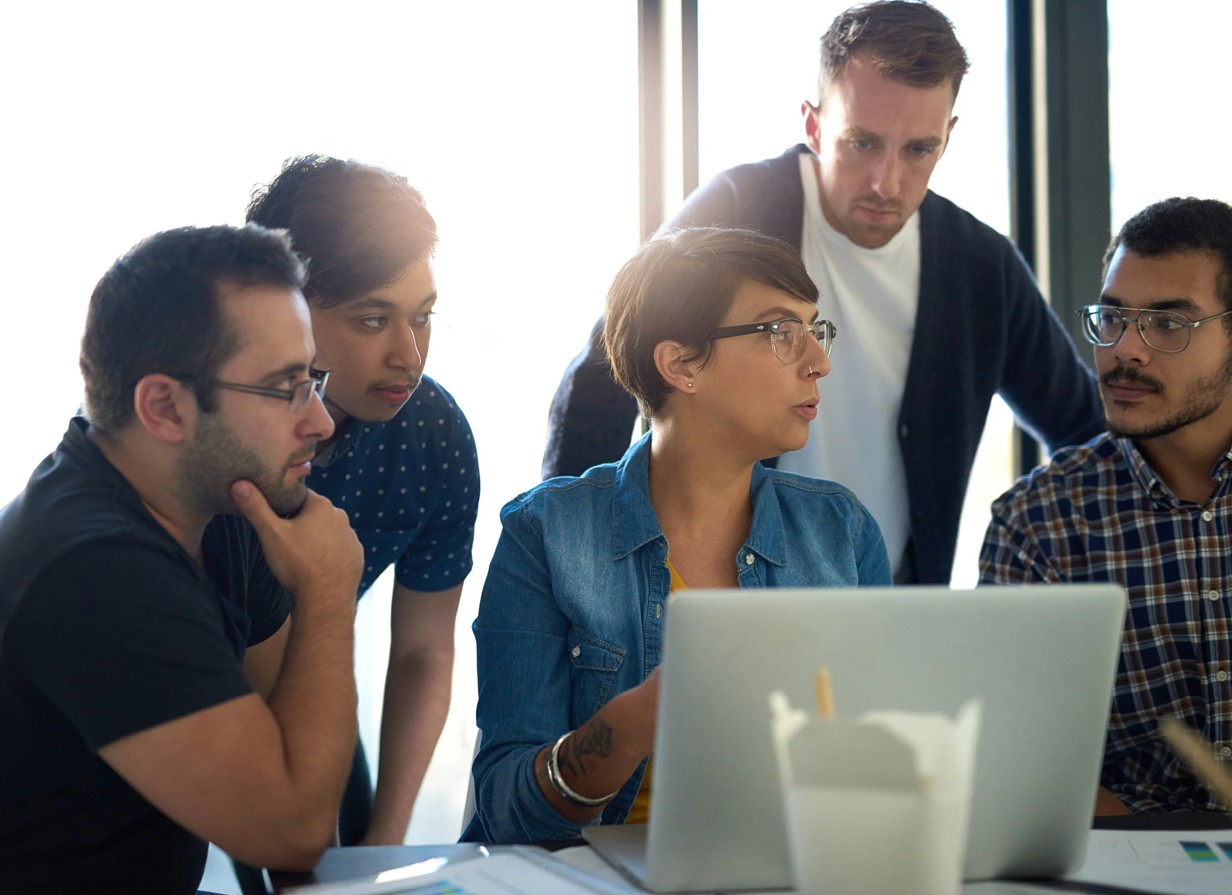 Five young adults in a meeting discussing around an open laptop in a bright room.